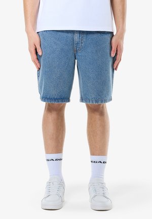 Homme portant un short en jean bleu mi-long, des chaussettes blanches avec du texte noir, des baskets blanches et une chemise blanche, debout les bras détendus le long du corps.