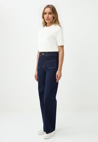 Jeans a gamba larga blu scuro con cuciture gialle a contrasto, vita alta e due tasche frontali. Indossati con un top bianco aderente a maniche corte.