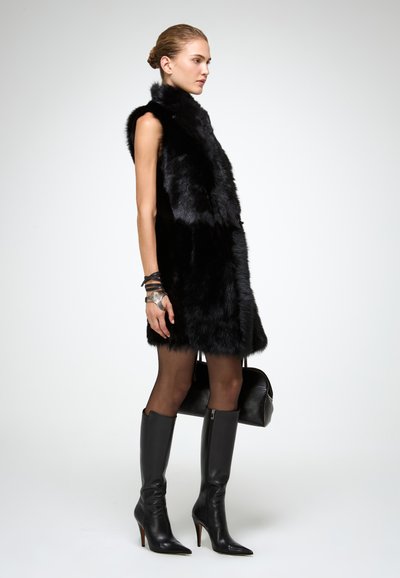 Juodas dirbtinis kailis liemenė su aukštu apykakle, be rankovių dizainas. Modelis dėvi juodas iki kelių odines batus, juodas raštuotas pėdkelnes ir laiko juodą rankinę.