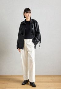Sort læderjakke med frynse detaljer, sort tætsiddende top, crème corduroy bukser og sorte hælede ankelstøvler på trægulv.