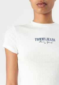 Haut blanc à manches courtes avec texture côtelée et petits motifs œillets, arborant un logo « TOMMY JEANS New York » en bleu marine sur le devant.