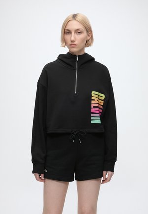 Ung kvinde med kort blondt hår iført en sort crop hoodie med farverig "CALIWIN" tekst og matchende sorte shorts.
