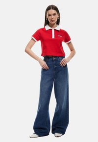 Rode cropped polo shirt met witte kraag en manchetten, gecombineerd met hoge taille, brede blauwe jeans. Model staat met handen op de heupen.