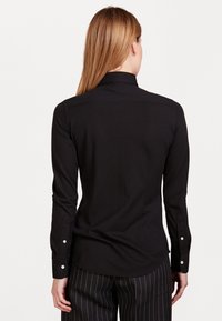 Black long-sleeve shirt with a collared neckline and buttoned cuffs. Smooth texture, fitted design, and rounded hem at the back.