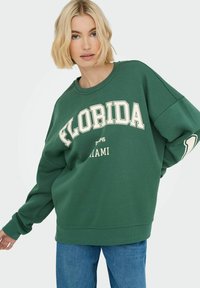 Felpa verde con la scritta "FLORIDA" e "MIAMI" in bianco. Vestibilità ampia con spalle cadenti e polsini a costine. Abbinata a jeans blu.