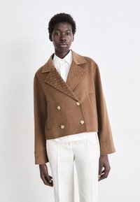Veste brune en laine, cropped, à double boutonnage avec de grands boutons dorés et un col texturé, portée par-dessus une chemise blanche à la silhouette structurée.
