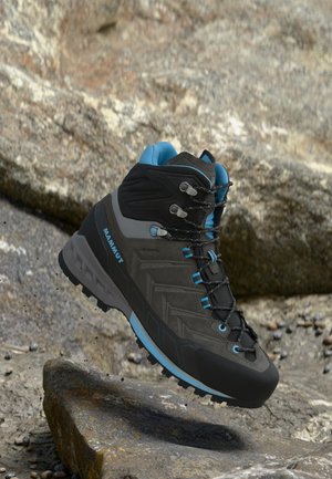 Bocanc de drumeție Mammut negru și albastru plutind deasupra unui teren stâncos, prezentând talpa detaliată și cârligele sigure pentru șireturi.