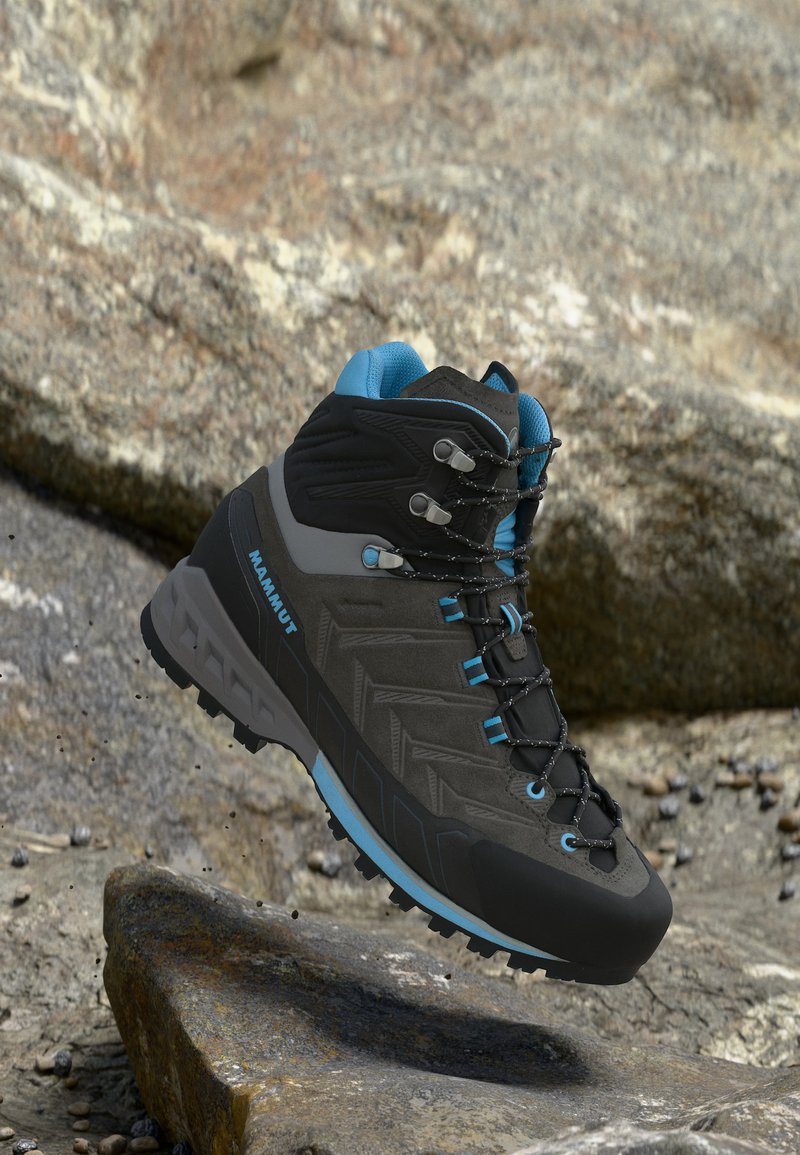 Černo-modrá turistická bota Mammut plovoucí nad kamenitou krajinou, zobrazující detailní vzorek podrážky a pevné háčky na tkaničky.