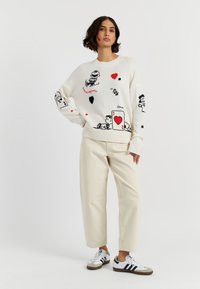 Witte gebreide trui met zwarte en rode cartoonafbeeldingen, met harten en figuren. Gecombineerd met beige wijde broek en zwartgestreepte schoenen.