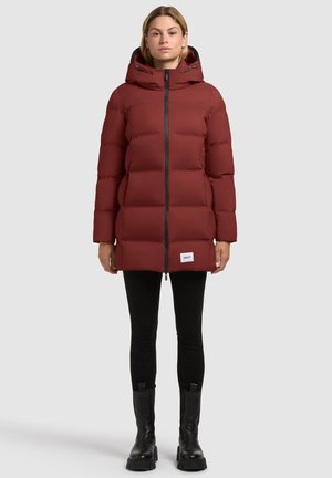 Eine Frau trägt einen langen, gepolsterten, rostroten Kapuzenmantel mit schwarzem Reißverschluss, schwarze Leggings und schwarze Stiefeletten und steht vor einem schlichten Hintergrund.