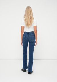 Laiendatud sinised teksad Denim materjalist, keskmise kõrgusega vöökoht ja kaks tagataskut. Kombineeritud lühikeste varrukatega valge särgiga ja mustade kingadega.