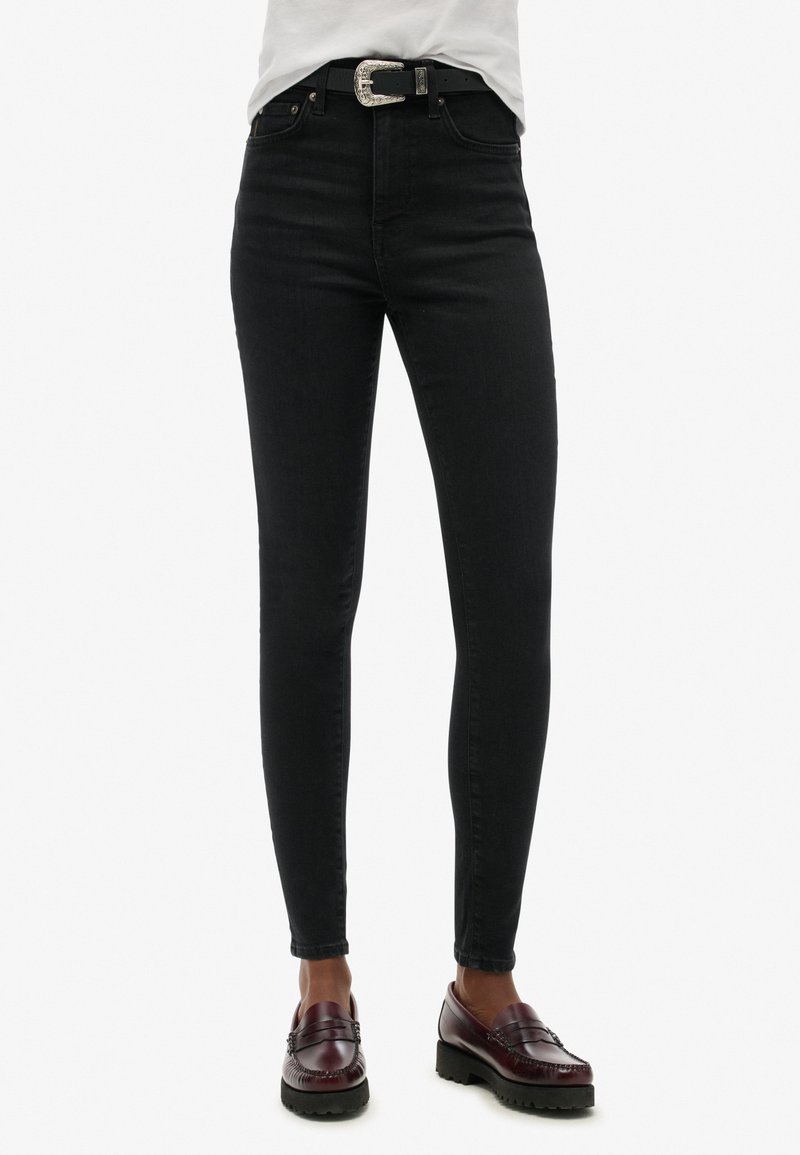 Zwarte high-waisted skinny jeans met een soepele textuur, zilverkleurige hardware op de zakken, gecombineerd met een zwarte riem en donkere instapschoenen.
