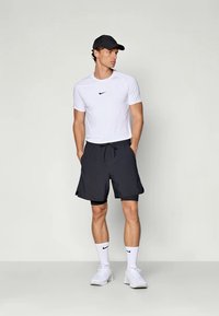 Mann mit schwarzer Mütze, weißem Nike T-Shirt, schwarzen mehreren Lagen Shorts, weißen Nike Socken und weißen Sneakers, der mit den Händen in den Taschen steht.