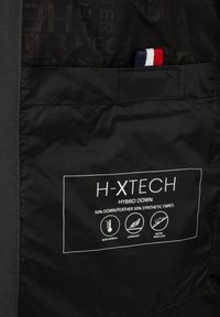 Notranjost črne izolirane jakne s oznako za nego, ki podrobno opisuje "H-XTECH hibridno puhovko." Sestava materiala: 50 % puha/plitvic, 50 % sintetičnih vlaken.