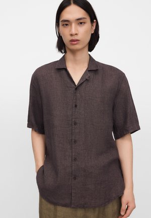 Młoda osoba o średniej długości czarnych włosach, ubrana w ciemnobrązową, krótkim rękawem koszulę zapinaną na guziki z kołnierzykiem, stoi z jedną ręką w kieszeni.