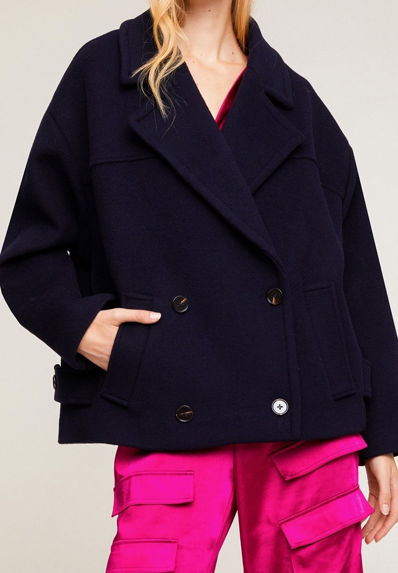 Manteau en mélange de laine bleu marine à double boutonnage avec de grands revers, poches latérales et boutons noirs, porté sur un pantalon cargo en satin rose vif.