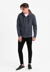Felpa con cappuccio e zip in pile grigio scuro, dotata di una fodera a quadri rossi, con lacci regolabili e tasche frontali. Indossata con jeans neri e sneakers.
