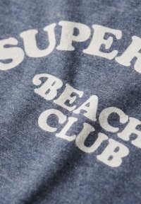 Tela azul jaspeada con grandes letras blancas que dicen "SUPER BEACH CLUB", con una apariencia suave y texturizada.
