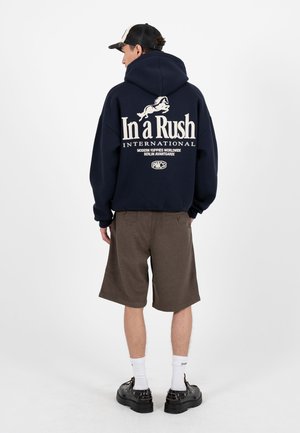 Homme portant un sweat à capuche noir avec du texte blanc et un motif de cheval, un short marron jusqu'aux genoux, des chaussettes blanches, des chaussures noires et une casquette noir et blanc, vu de dos.