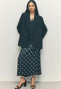 Blazer noir à double boutonnage sur un haut vert foncé, associé à une jupe à pois bleu canard et des sandales à talons noires. Tissu lisse et coupe élégante.