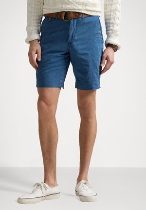 Pantalones cortos azules texturizados con un frente plano, bolsillos laterales y un cinturón; combinados con zapatos blancos con cordones y un suéter de punto color crema.