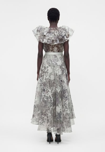 Γυναίκα που φοράει διάφανο, πολύστιχο φόρεμα με λουλουδάτο σχέδιο, με βολάν στους ώμους και εφαρμοστή μέση, συνδυασμένο με μαύρα ψηλοτάκουνα.