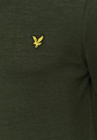 Pull en maille vert foncé avec un petit logo d'aigle brodé jaune sur la poitrine. La texture semble lisse et ajustée.