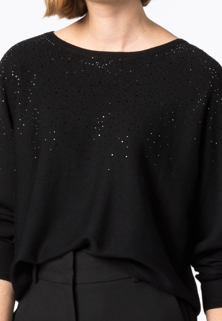 Pull noir à manches longues avec de petites décorations scintillantes dispersées sur le haut de la poitrine et les épaules, porté avec un pantalon noir.