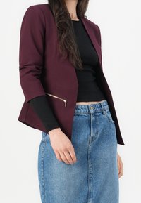 Blazer sur mesure bordeaux à coupe ajustée, détail de poche zippée, associé à un haut noir et une jupe en jean taille haute.