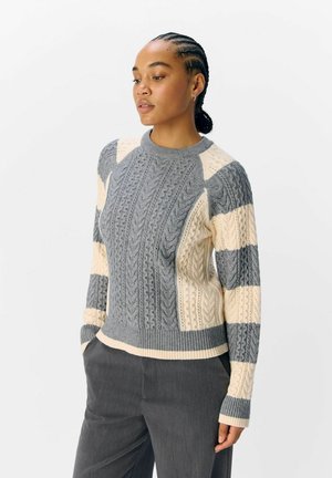 Donna con capelli intrecciati, che indossa un maglione grigio e crema a trecce e pantaloni scuri, in piedi con le mani in tasca, guardando in basso e di lato.