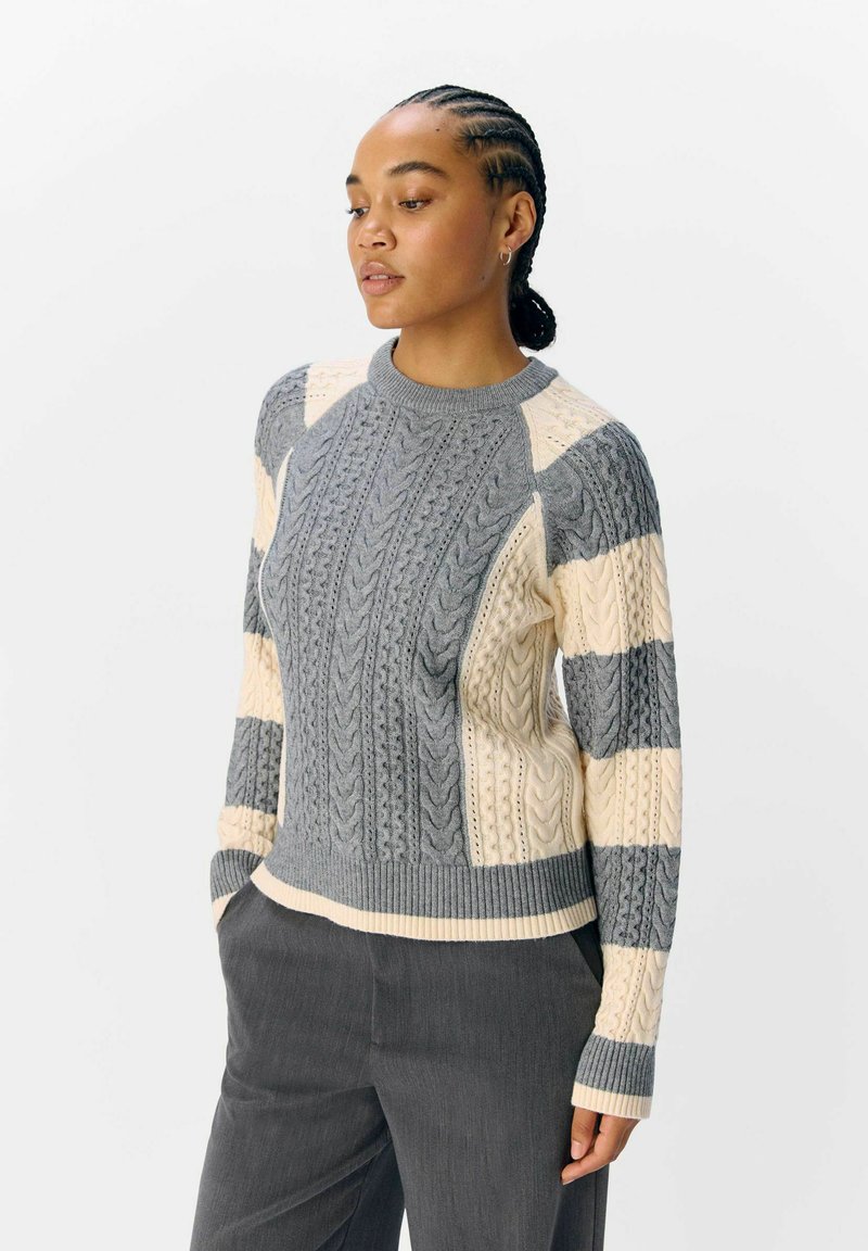 Vrouw met vlechten die een grijs en crème kabelgebreid sweater en donkere broek draagt, met de handen in de zakken, kijkend naar beneden en naar de zijkant.