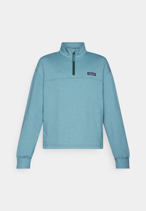 Šviesiai melsvas vilnonis megztinis su aukštu apykakle ir pusiau užtrauktuku. Jame yra horizontalus siūlas ir mažas logotipas ant krūtinės. Minkšta tekstūra.