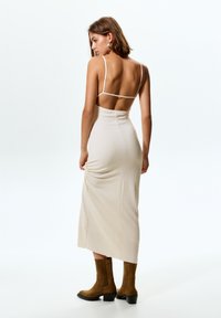 Bež oblečena obleka s tankimi naramnicami, teksturirana tkanina in razporek do stegna; v kombinaciji z rjavimi škornji do kolen in minimalističnimi oblikovalskimi akcenti.