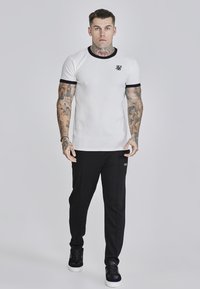 Witte overhemd met korte mouwen en zwarte kraag en manchetten, met een klein logo. Zwarte broek met een slim fit en zijaccenten. Zwarte sneakers.