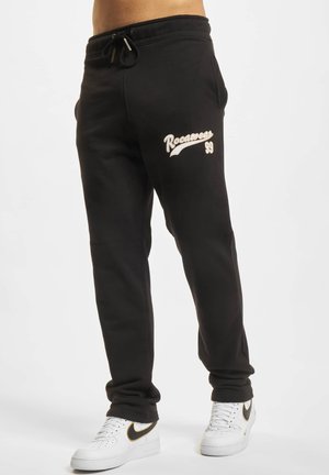 Sort sweatpants lavet af bomuldsblanding, med snor i taljen og "Rocawear 99" logo i beige på venstre lår. Samlet med hvide sneakers.