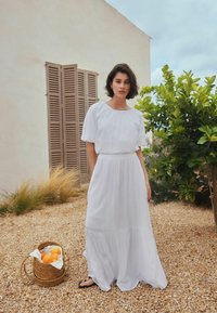 Vestido maxi blanco con mangas cortas abullonadas y bajo de volantes. Tela suave, ajuste relajado. Cesta tejida con naranjas en primer plano.