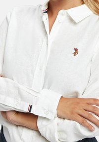 Vit linneskjorta med klassisk krage, knäppning fram och broderad logotyp på bröstet. Manschetterna har röd, vit och blå randig accent.