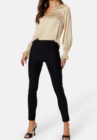 Haut en satin crème avec un col en V et des manches ballon, associé à un pantalon noir ajusté et des talons noirs à lanières. Accents texturés sur les manches visibles.