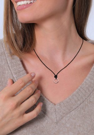 Black cord necklace with a silver ring pendant, worn against a light beige sweater with a V-neck design. Smooth texture, minimalistic design.