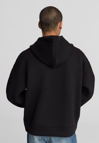 Felpa nera con un fit rilassato, caratterizzata da un ampio cappuccio e spalle scese. Il tessuto appare morbido, con una texture liscia.