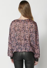 Durchsichtige Bluse mit langen Ärmeln im Paisleymuster in Erdtonen und blauen Akzenten. Schwarze strukturierte Manschetten und Knopfverschluss hinten.