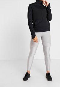 Sort lynlåstrøje med høj krave, kombineret med lysegrå leggings med teksturerede mønstre og en lille logo. Sorte sportssko.