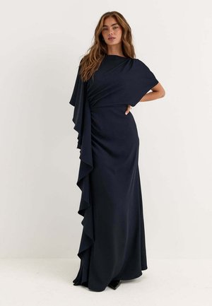 Donna con lunghi capelli castani che indossa un abito nero lungo fino a terra con un dettaglio di balze drappeggiate su un lato, su uno sfondo chiaro e uniforme.