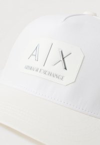 Bijela bejzbol kapa s bijelim zakrpom koja ima sjajni srebrni logo "A|X" i oznaku "Armani Exchange" na prednjem dijelu.