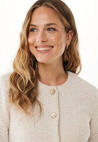 Textured crèmekleurige cardigan met ronde halslijn, voorzien van twee gouden decoratieve knopen en een iets boxy ontwerp. Glad haar valt naar beneden.
