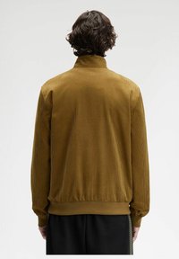 Una persona con capelli ricci sta in piedi di spalle, indossando un giubbotto in velluto a coste giallo senape e pantaloni neri con una striscia decorata di lato.