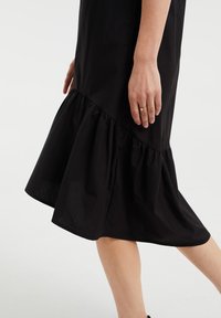 Schwarzes Kleid mit ausgestelltem Saum und Rüschendetail, aus leichtem Stoff, der bis knapp über das Knöchel reicht. Eine Hand ist seitlich sichtbar.