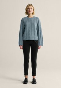 Vaaleansininen palmikkoneulepusero leveillä hihoilla, yhdistetty mustiin kapeisiin housuihin ja mustiin loafereihin. Sileä pinta ja rento istuvuus.