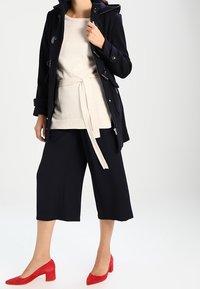 Marineblauer Mantel mit Kapuze, weißer strukturierter Pullover mit Bindegürtel und schwarze Culottes. Rote spitze Absatzschuhe runden das Outfit ab.