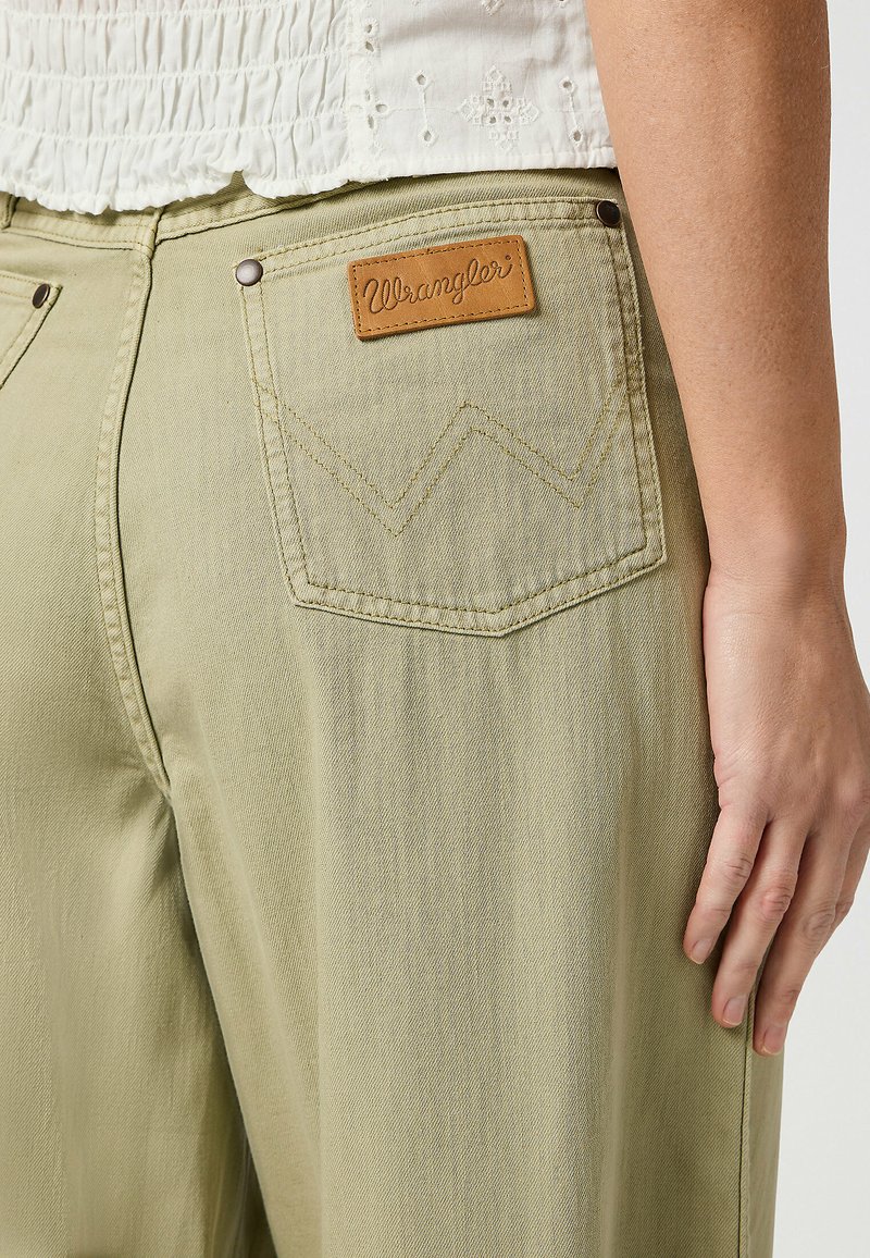 Un jean Wrangler vert clair présente une étiquette en cuir sur la poche arrière avec le logo de la marque et un détail de couture en zigzag. Texture lisse.
